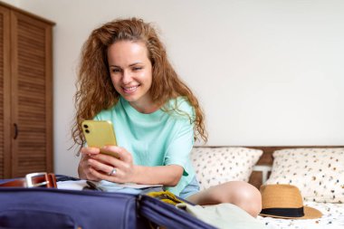 Heyecanlı kıvırcık kadın, yatağında çantasıyla otururken telefonunu kullanarak internetten otel rezervasyonu yapıyor..