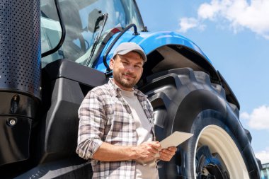 İnsan tarım makineleri tamircisi traktörün yanında duruyor ve elinde dijital tabletle kameraya bakıyor ve gülümsüyor..