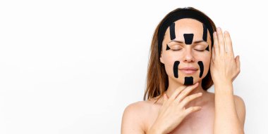 Güzellik mankeni, beyaz arkaplan önünde yüzüne Kinesio bantları yapıştıran esmer bir kadın. Güzellik, vücut ve cilt bakımı.