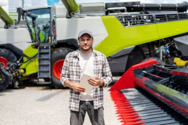 Tarım makineleri satma veya satın alma kavramı. Dijital tabletli bir adam bir galeride hasatçıların önünde duruyor..