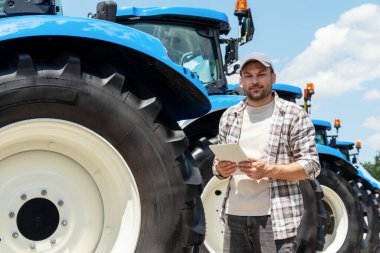 Tarım makineleri satma veya satın alma kavramı. Dijital tabletli bir adam yeni çiftlik traktörlerinin önünde duruyor..