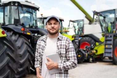 Elinde tablet olan bir adamın portresi bir bayilikte yeni tarım makinelerinin önünde duruyor..