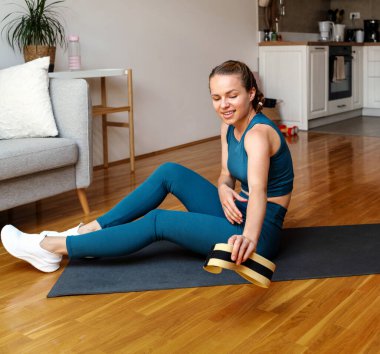 Evde idman yapıyorum. Kadın elinde küçük bir direniş bandıyla egzersiz minderinde oturuyor..