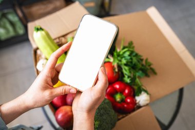 Olgun sebzeler ve meyvelerle dolu kutunun üstündeki kadın ellerinde akıllı telefonun sahte ekranı. Çiftlik sebzeleri ve meyve mobil teslimat hizmeti.