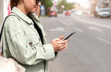 Kadın şehir yolunun yanında duruyor ve akıllı telefonunu internet taksi rezervasyonu ya da araba paylaşım servisi için kullanıyor.. 