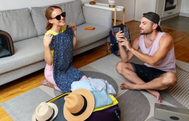 Couple sits on the floor at home packing or unpacking  travel bags trying on a dress, man photographing woman while she trying on a dress. clipart