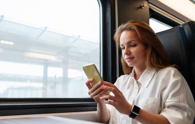 Modern teknoloji meraklısı iş kadını iş için trenle seyahat ederken akıllı telefonuyla internetten mesajlaşıyor. Devam et..