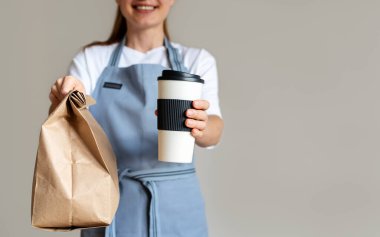 Kahve ve paket yemek. Önlüklü bir kadının elinde içinde yiyecek ve büyük bir fincan kahve olan bir kese kağıdı..