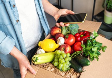 İnternetten sebze ve meyve yetiştiriyorlar. Kadın eli boş cep telefonu ve taze sebzeli karton kutu..