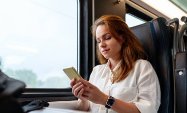 Kafkasyalı genç iş kadını trenle seyahat ederken akıllı telefonunu kullanarak çevrimiçi mesajlaşıyor..