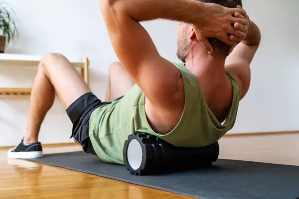 Erkek sporcu spor yapıyor. Köpük silindiri kullanıyor, evde egzersiz yapıyor, vücut bakımı yapıyor..