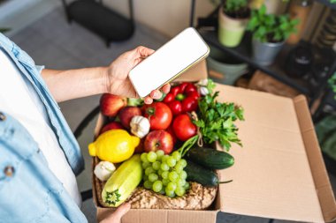 Mobil uygulama çiftlik sebze siparişi veriyor. Cep telefonunda boş ekran var. Kutunun üstünde taze ve olgun organik sebzeler var..