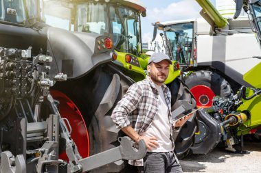 Elinde dijital tablet olan beyaz bir adam bir malzeme satıcısının yeni tarım makinesinin yanında duruyor..
