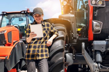 Tarım ve inşaat malzemeleri satıcısı. Ekose gömlekli ve şapkalı kadın yeni traktörün direksiyonuna yaslandı ve dijital tablet kullandı..
