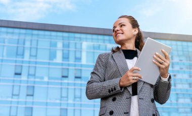 Modern ofis binasının önünde duran tabletli kendine güvenen bir iş kadını..