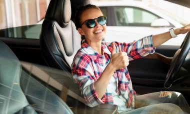 Genç bir dişi galeride yeni arabasında oturuyor, gülümsüyor ve işaret veriyor. Araba kiralama servisi, araba paylaşımı..