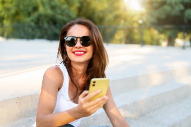 Dışarıdaki merdivenlerde oturan ve cep telefonunu kullanan güneş gözlüklü kadın..