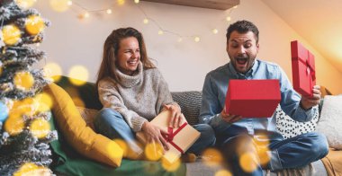 Mutlu çift evdeki koltukta Noel hediyelerini açıyor. Kadın gülerken erkek sürpriz bir hediye açar..