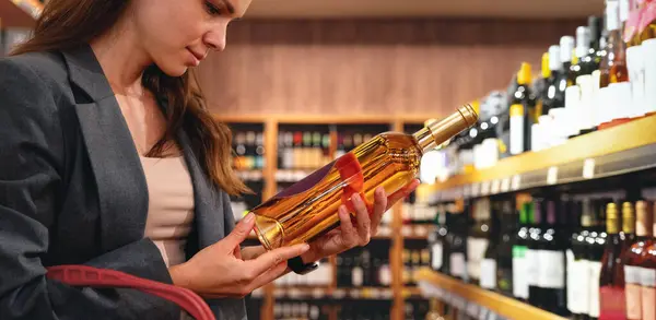 Mağazada şarap etiketi okuyan bir kadın, seçimini dikkatlice yapıyor..
