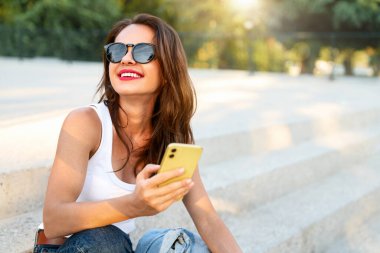 Sıradan kıyafetli şehir kadını merdivenlerde oturuyor, akıllı telefon kullanırken gülümsüyor..