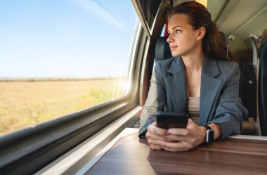 Akıllı telefonlu bir iş kadını, trenle gidip gelirken pencereden dışarı bakıyor. İş seyahati kavramı. 