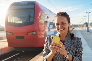 Kadın banliyö treni bir demiryolu platformunda duruyor ve arka planda bulanık bir trenle akıllı telefonunu kullanarak gülümsüyor. Online tren biletleri rezervasyonu. 