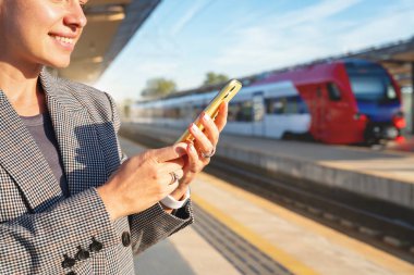 İnternetten tren bileti rezervasyonu. Kadın banliyö bir demiryolu platformunda duruyor, arka planda bulanık bir trenle akıllı telefonunu kullanıyor..