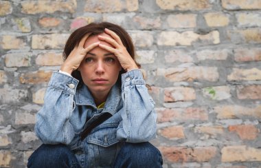 Yorgun ve endişeli bir yüzü olan kadın dışarıda tuğla duvarın yanında oturuyor, elleri alnında..