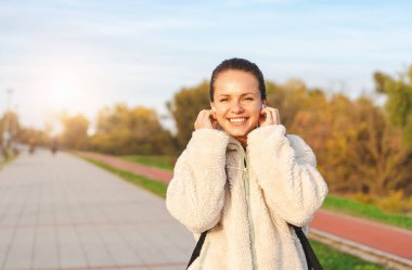 Bej ceketli ve kulaklıklı neşeli beyaz kadın parkta yürürken kameraya bakıyor ve gülümsüyor..
