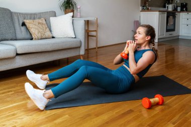 Yoga minderinde oturan motive olmuş bir kadın ev egzersizi sırasında bir halterle karın egzersizi yapıyor..