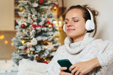 Beyaz kadın portresi, akıllı telefon ve kablosuz kulaklıkla koltukta oturuyor, Noel tatillerinde müzik dinliyor..