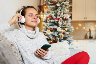 Kanepede oturan akıllı telefon ve kablosuz kulaklıklarla Noel tatili boyunca müzik dinleyen bir kadın..