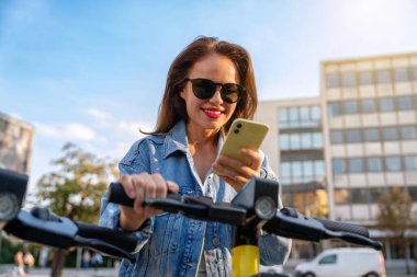 Kadın şehir sakini şehrin caddesinde akıllı telefonuyla elektrikli scooterın kilidini açıyor..