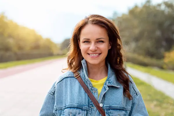 Kot ceketli, dışarıda yürüyen, kameraya bakıp gülümseyen esmer beyaz kadın portresi..