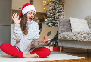 Noel arifesinde evde dizüstü bilgisayar kullanan ve ekranda el sallayan Noel Baba şapkalı kadın..