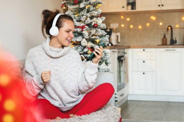 Kablosuz kulaklıklı bir kadın evdeki koltukta oturuyor ve akıllı telefonunu kullanıyor, Noel tatilinde müzik dinliyor..