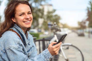 Bir şehir caddesinde bankta oturan akıllı telefonlu gülümseyen şehirli kadın..