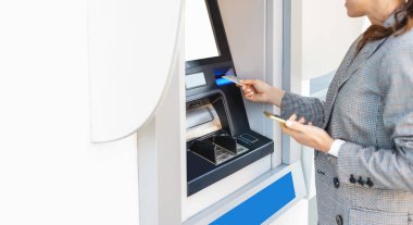 Şehir caddesinde akıllı telefonu ve kredi kartı olan bir ATM kullanan şık giysili bir iş kadını.