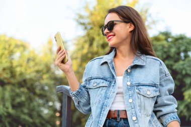 Kot ceketli ve güneş gözlüklü bir kadın dışarıda akıllı telefon kullanıyor, arka planda bulanık yeşillik var..