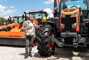 Dijital tabletli beyaz bir adam açık havada, bayilikte inşaat ve tarım makinelerinin yanında duruyor..