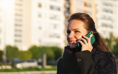 Siyah ceketli ve fularlı bir kadın şehir caddesinde cep telefonuyla konuşuyor..