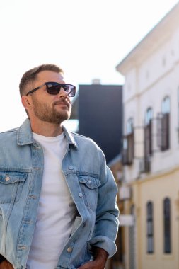 Şehirde güneş gözlüğü ve kot ceketli beyaz bir adam yürüyor..