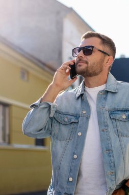 Güneş gözlüklü beyaz adam şehir caddesinde telefonla konuşuyor..