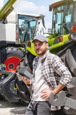 Tarımsal ekipman bayisinde dijital tabletle traktöre karşı çalışan erkek tarım işçisi.