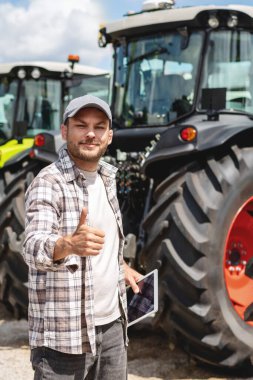 Dikey portre erkek çiftçi bir sonraki çiftlik traktörleri ve jestler baş parmak yukarı.