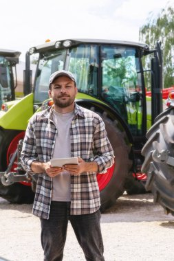 Tarımsal ekipman bayiliğinde traktörlerin arka planında dijital tablet tutan beyaz bir adam..