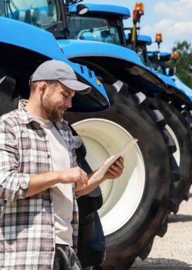 Tarım makinelerinde yeni traktörlerin yanında dijital tablet kullanan ve gülümseyen erkek çiftçi..