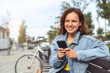 Neşeli Kafkasyalı kadın şehir caddesinde bir bankta oturuyor, cep telefonunu kullanıyor ve gülümsüyor..