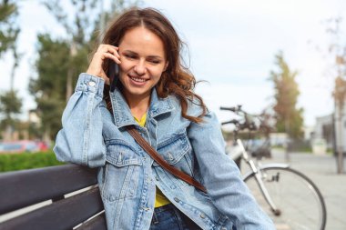 Kot ceketli genç şehir kadını sokakta otururken telefonla konuşuyor..