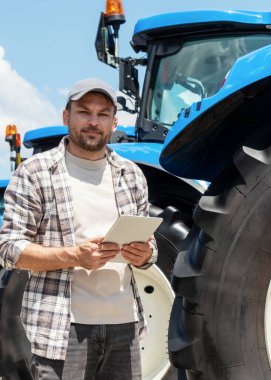 Dijital tabletli adam yeni çiftlik traktörünün yanında duruyor.